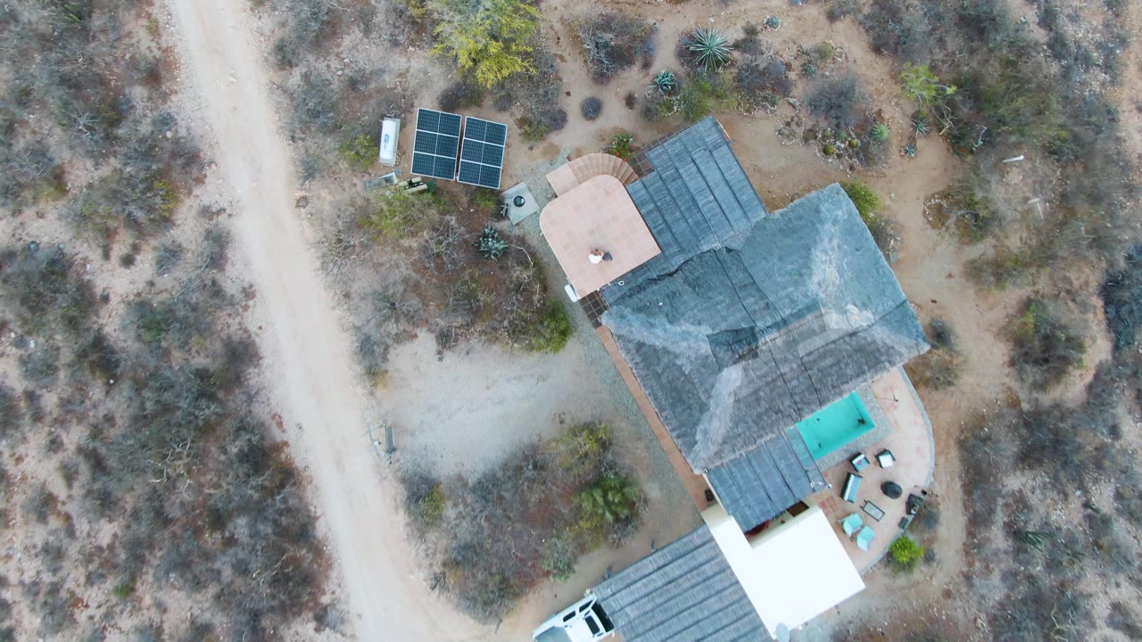 Aerial frame in Cabo Pulmo, Baja California Sur showing the house, solar panels, small pool, patio area, white vehicle near the lower edge, dirt roads, and no visible people.