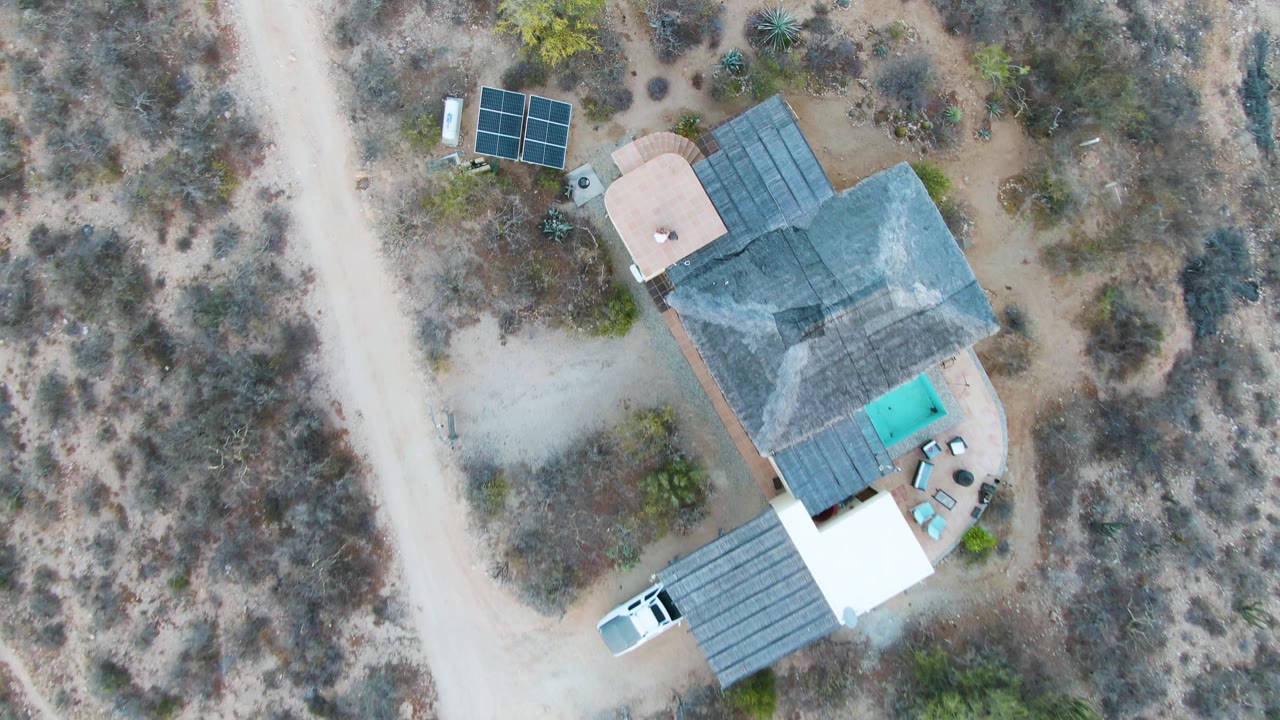Top-down frame from Cabo Pulmo, Baja California Sur with the house shifted toward the upper right, a white vehicle on the lower dirt track, solar panels, dry brush, and no visible people.