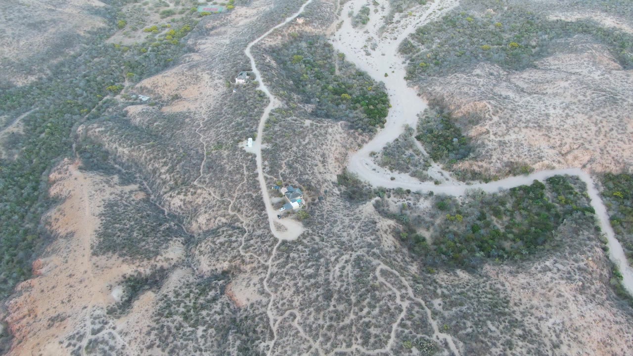 Overhead drone view of a winding dirt road crossing dry scrub-covered hills near Cabo Pulmo, Baja California Sur, with a small structure and no visible people.