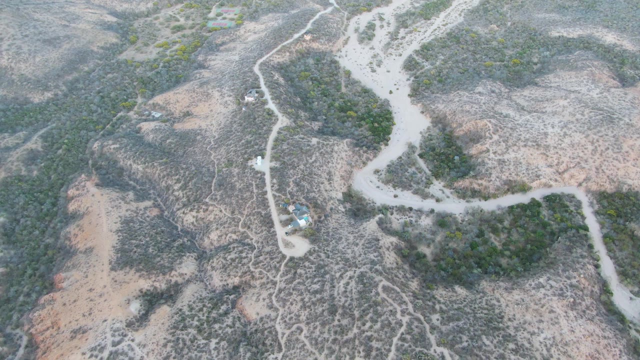 High aerial frame over Cabo Pulmo backcountry showing a pale dirt road crossing dry rocky hills with green scrub, a small vehicle on the track, and no visible people.
