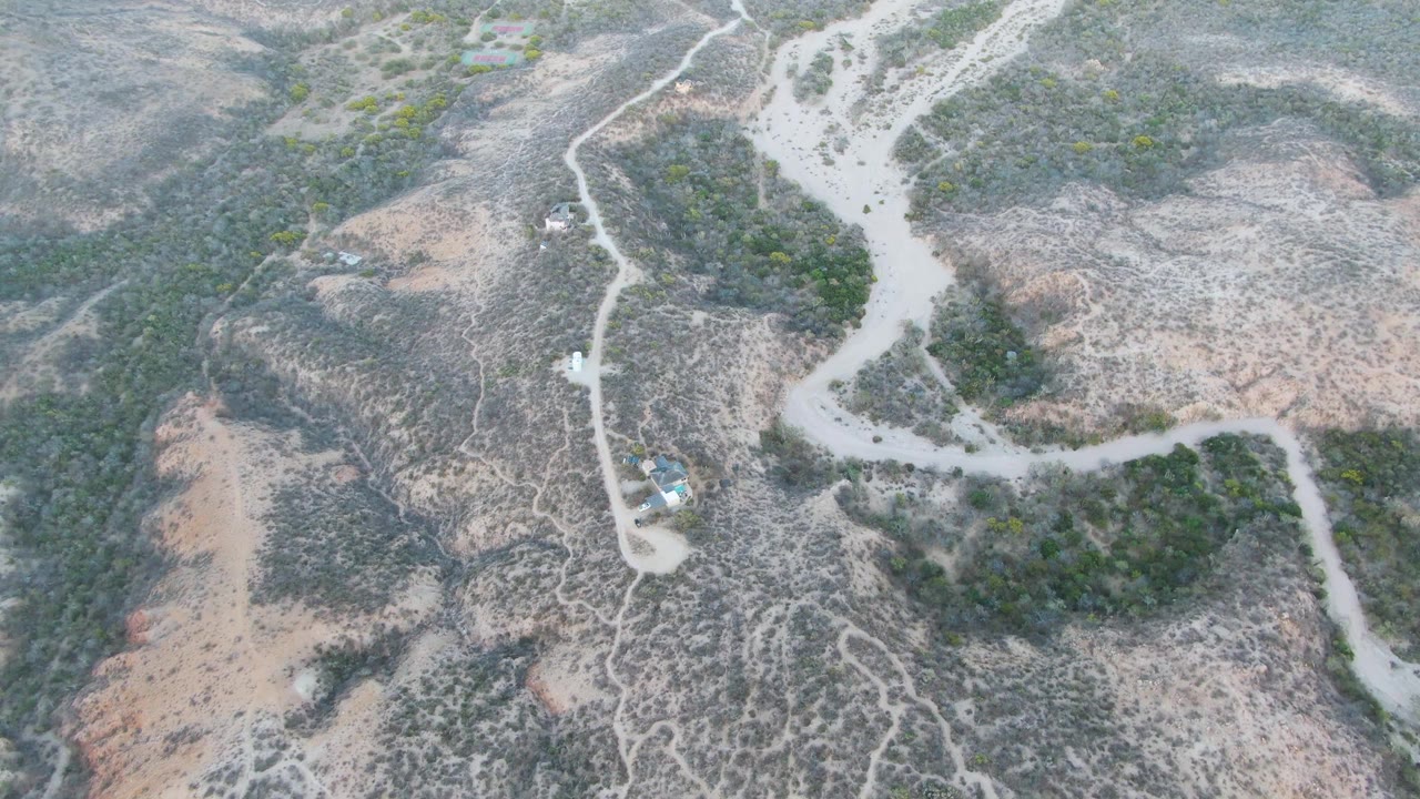 Drone view of a vehicle on a winding dirt path in arid scrubland.