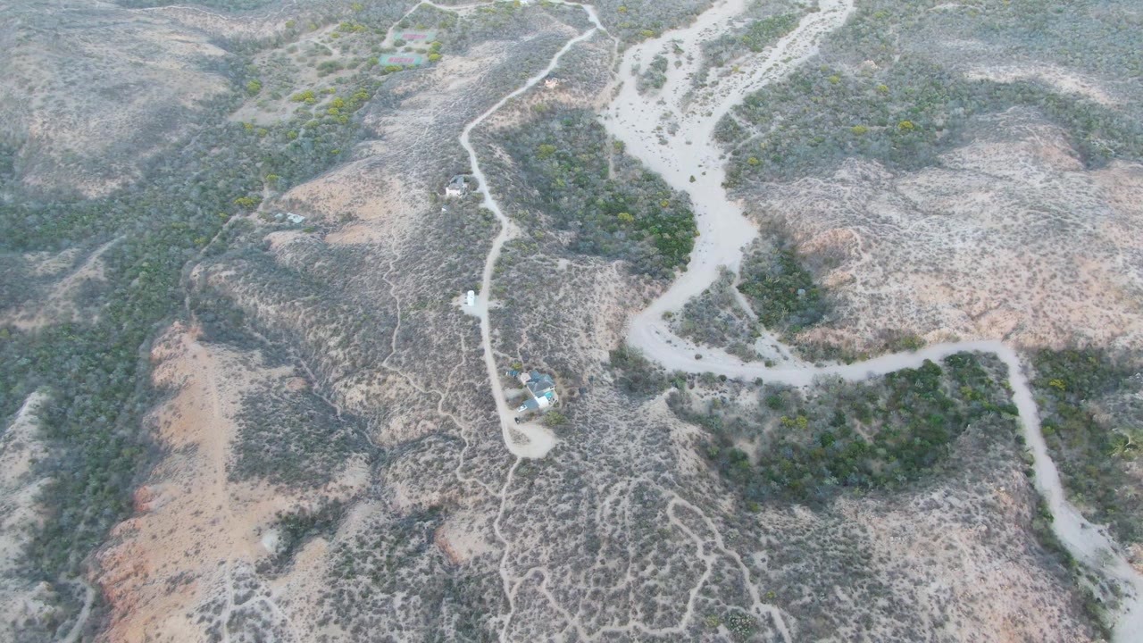 Aerial frame over Cabo Pulmo dry terrain showing a small vehicle on a winding pale dirt road between rocky slopes and green scrub, with no visible people.