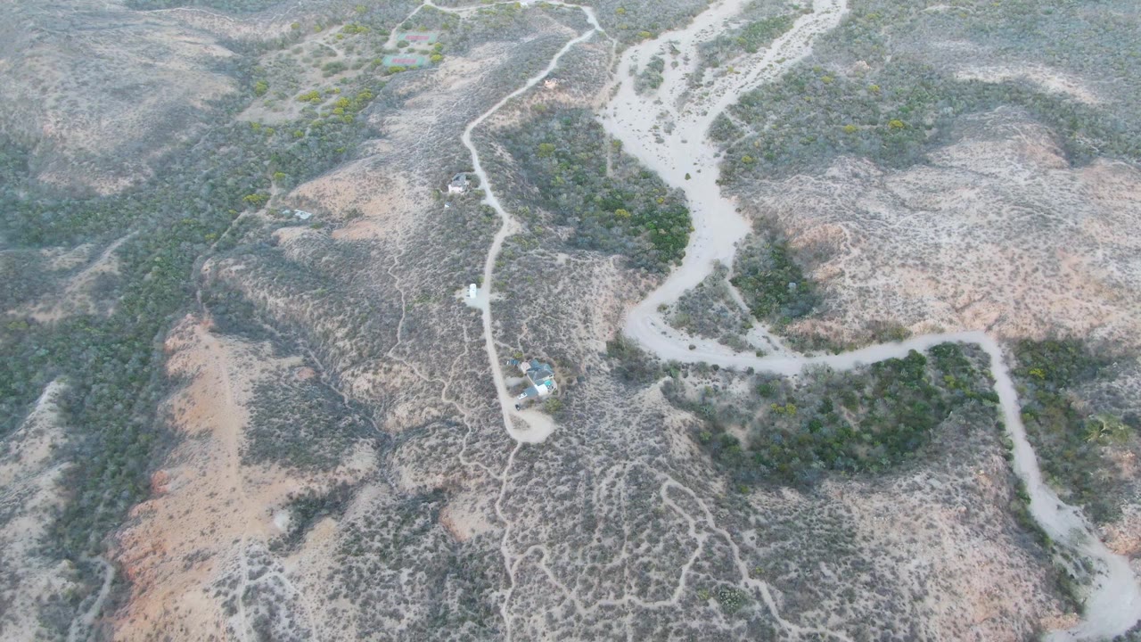 The frame looks down on Cabo Pulmo hills where a pale dirt road bends around scrubby green patches and a small vehicle remains visible, with no visible people.