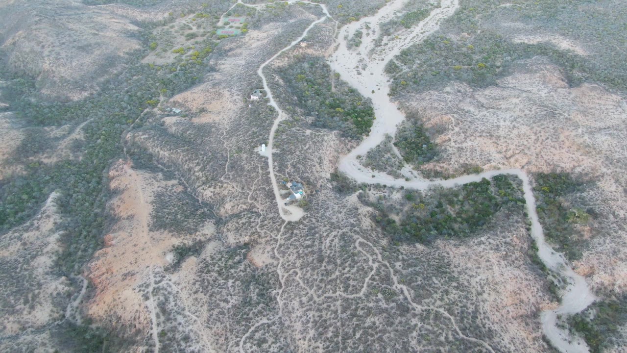 Aerial view in Cabo Pulmo showing the car slightly farther along the pale road, with scrubby green patches and dry ridges filling the frame, no visible people.