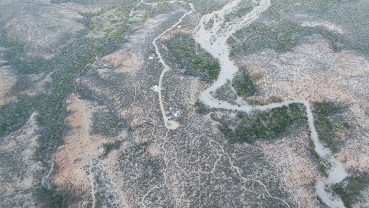 High aerial frame over Cabo Pulmo terrain with the vehicle on a curving dirt track beside low green vegetation and exposed dry ground, no visible people.