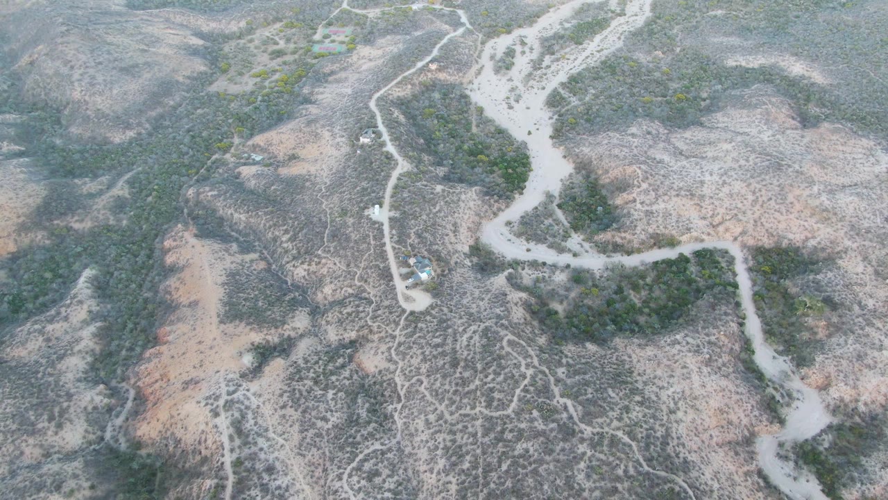 Cabo Pulmo aerial frame with the car on the pale road near a cluster of darker green scrub, surrounded by gray and tan dry hillside, no visible people.