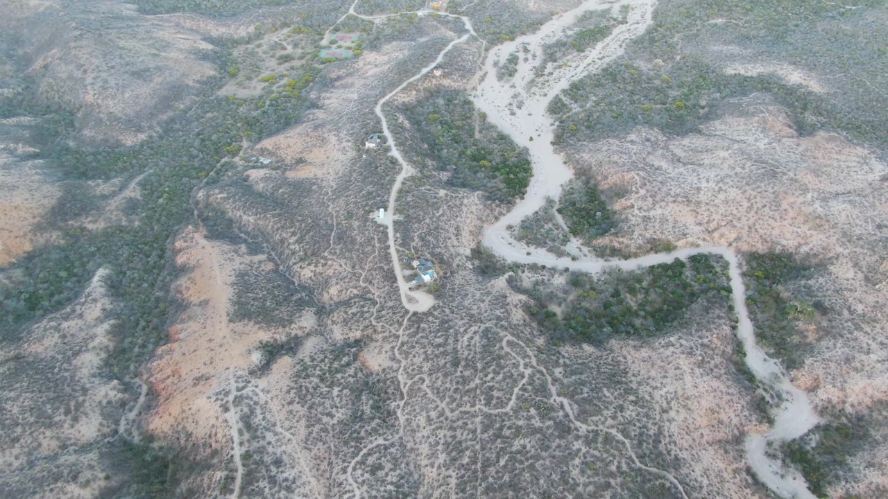 Aerial frame from Cabo Pulmo, Baja California Sur showing a pale dirt road crossing dry hills, sparse green brush, a small vehicle on the road, and no visible people.