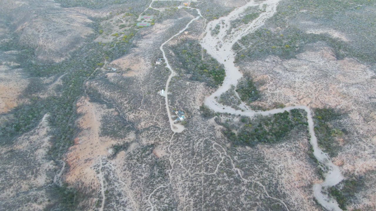 High aerial frame over Cabo Pulmo, Baja California Sur showing the road branching and curving through arid hills, with the small car near the route and no visible people.
