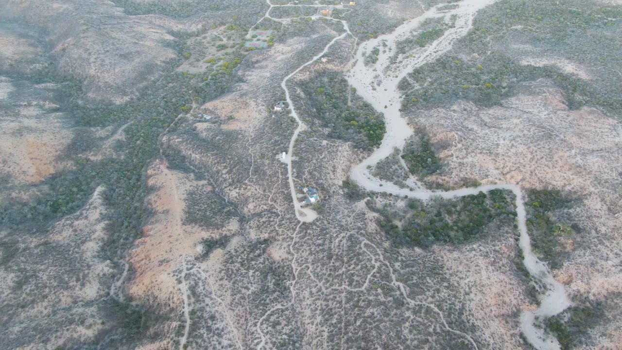 High aerial frame over Cabo Pulmo arid hills shows a small light-colored vehicle on a winding pale dirt path beside a dry riverbed, with sparse green vegetation and no visible people.