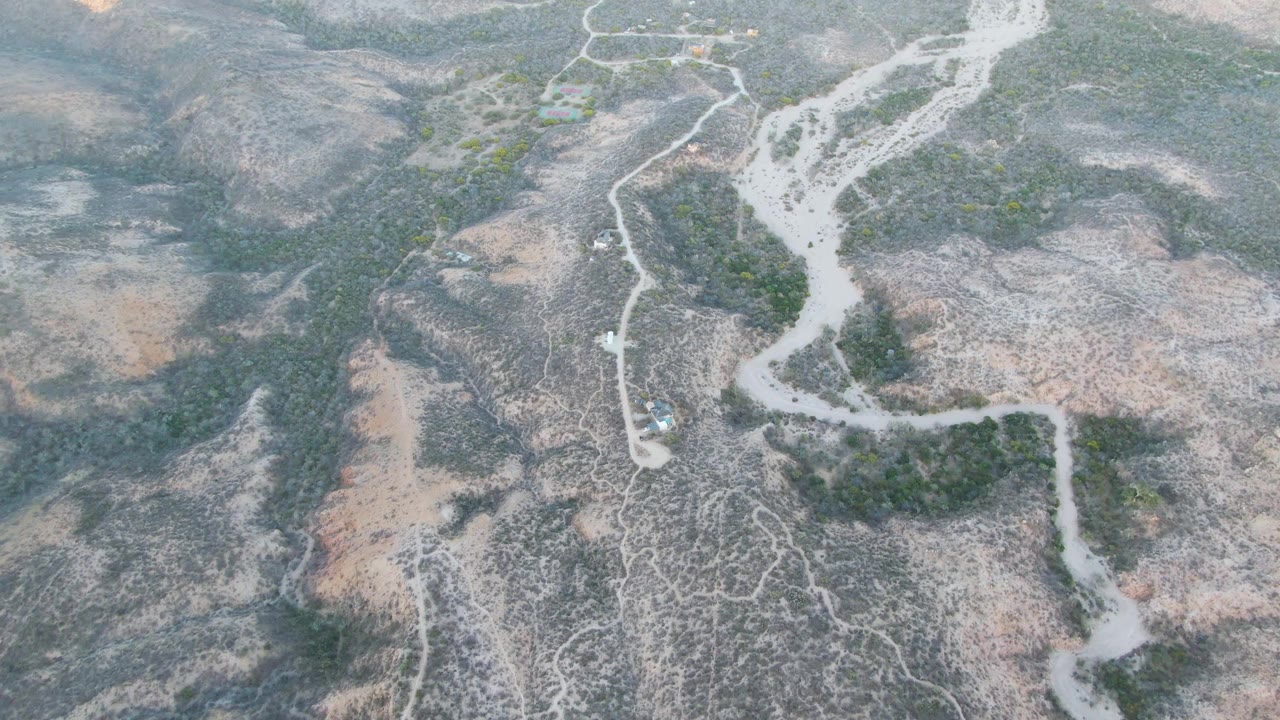 High aerial view near Cabo Pulmo, Baja California Sur, with the exposed riverbed centered between brown slopes, faint dirt tracks, scattered shrubs, and no visible people.