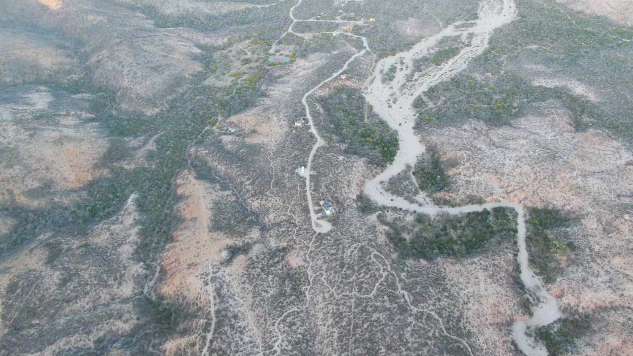 High aerial frame over Cabo Pulmo desert terrain showing a pale winding road, scattered green vegetation, dry washes, and no visible people at the start of the clip.