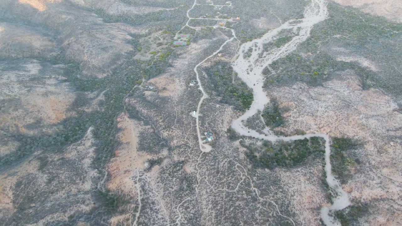 High aerial frame over Cabo Pulmo, Baja California Sur, showing a pale dry riverbed through rocky arid hills with sparse green vegetation and no visible people.
