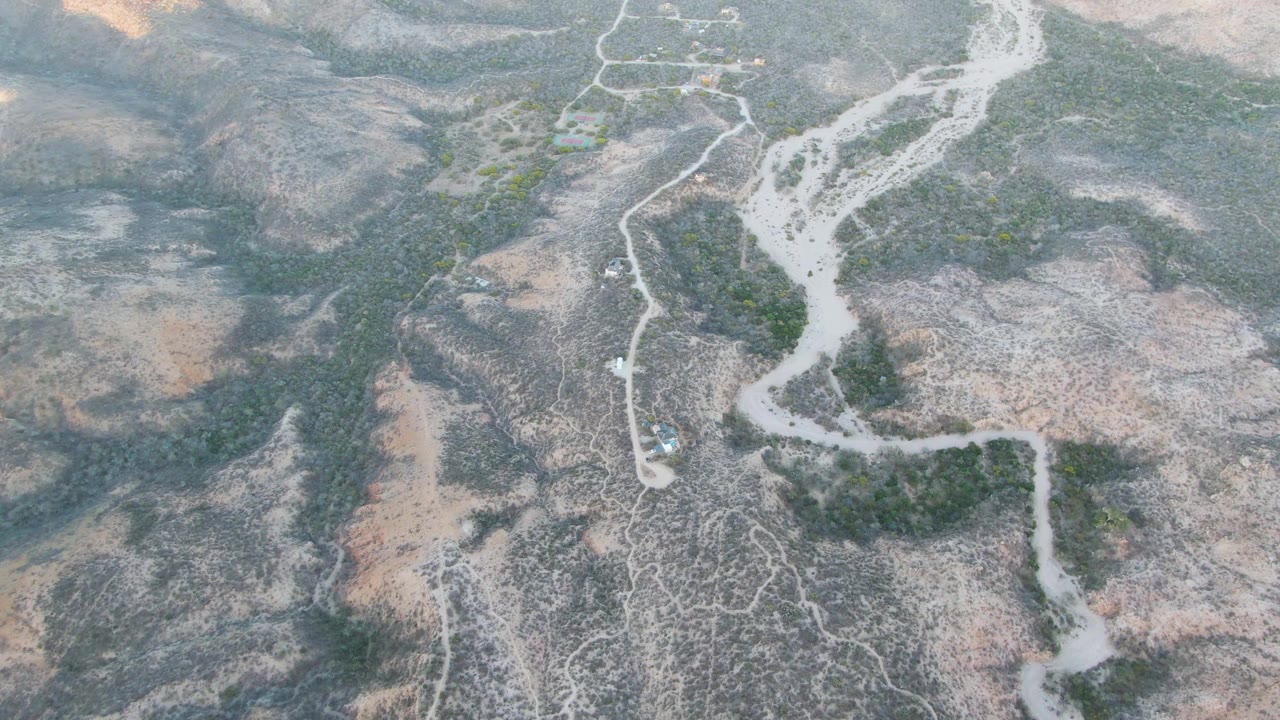 Aerial view in Cabo Pulmo with the white dry riverbed running down the center, branching dirt paths nearby, muted rocky slopes, and no visible people.