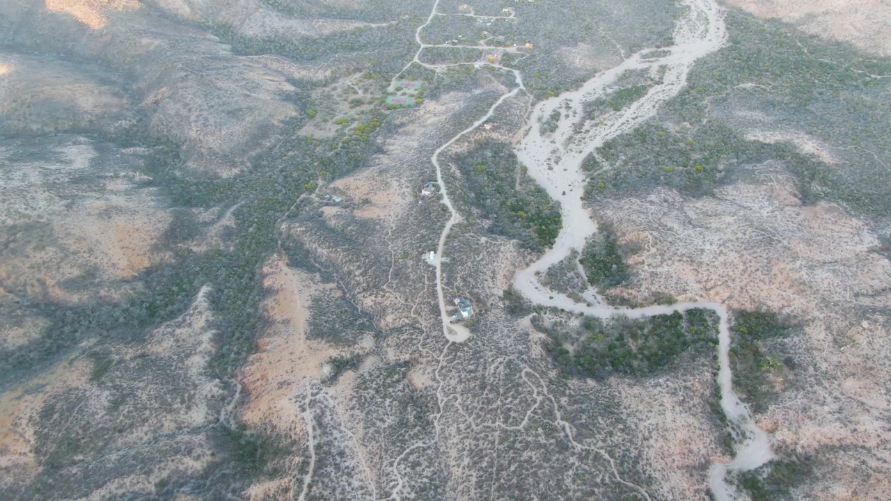 High drone-style frame over Cabo Pulmo showing a winding pale riverbed, rough dirt tracks, scrub patches, and a small light vehicle near the wash with no visible people.