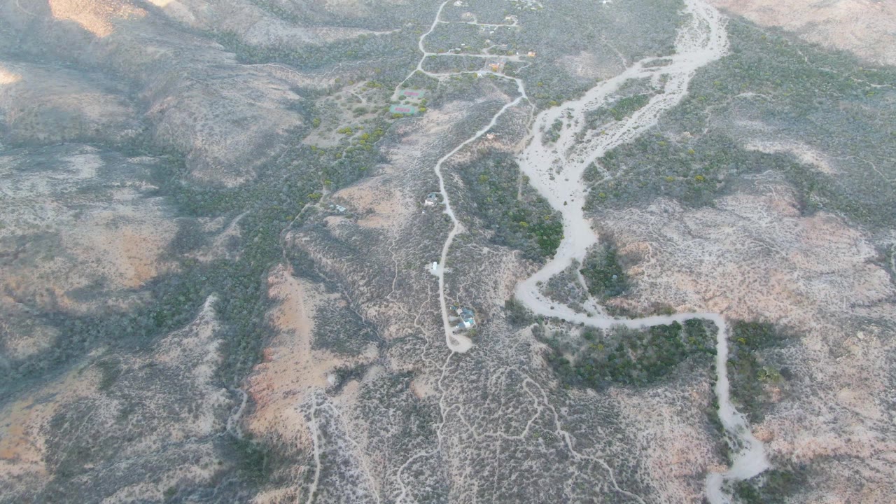 Overhead drone view near Cabo Pulmo, Baja California Sur, showing a pale dry riverbed crossing rocky brown ground with sparse green vegetation and no visible people.