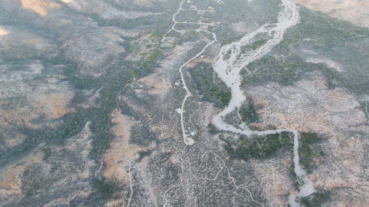 Overhead view near Cabo Pulmo, Baja California Sur, showing a pale dry channel splitting through gray-brown rocky land, small green patches, faint tracks, and no visible people.