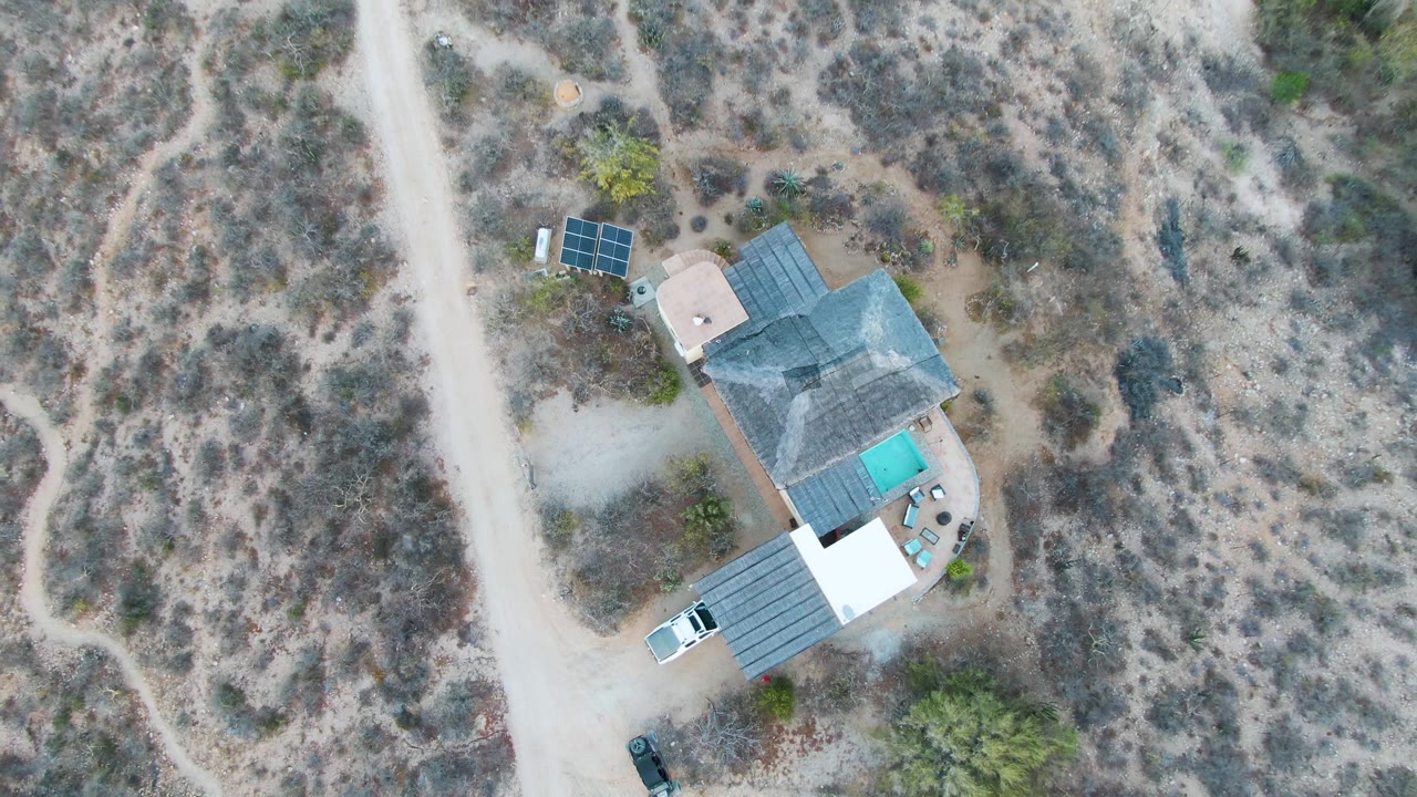 Opening overhead frame of a house in Cabo Pulmo, Baja California Sur, with roof solar panels, a small pool, a dirt driveway, parked vehicles, dry scrubland, and no visible people.