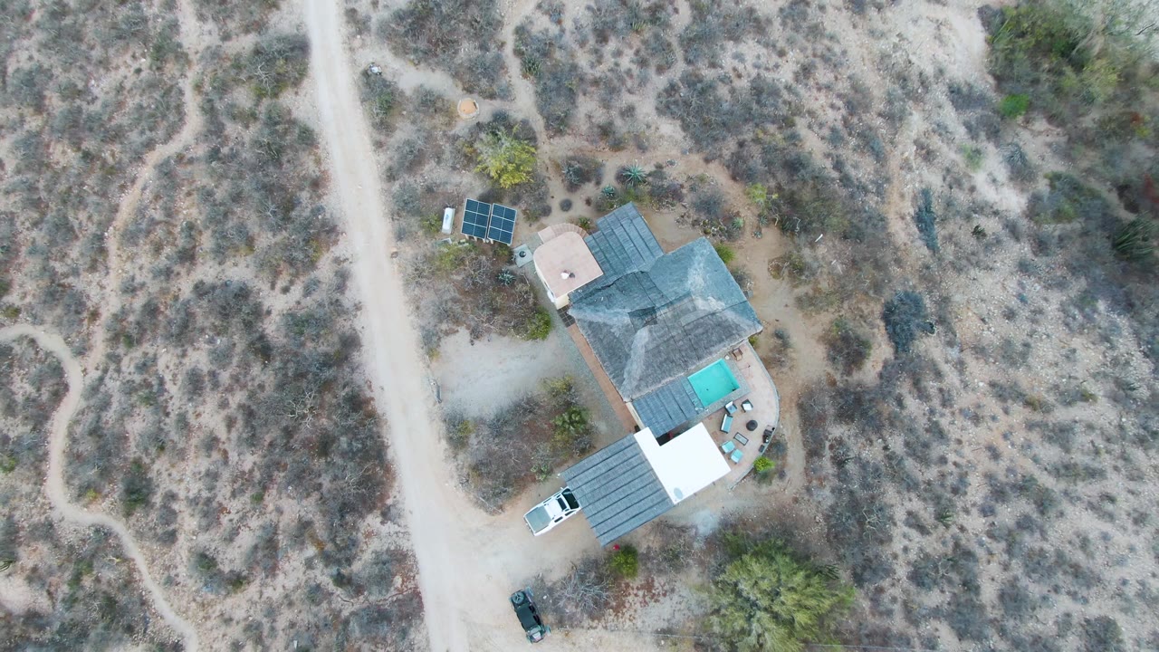 One second into the clip, the Cabo Pulmo house remains near the left of frame with its pool, solar panels, dirt access road, two parked vehicles, surrounding scrub, and no visible people.