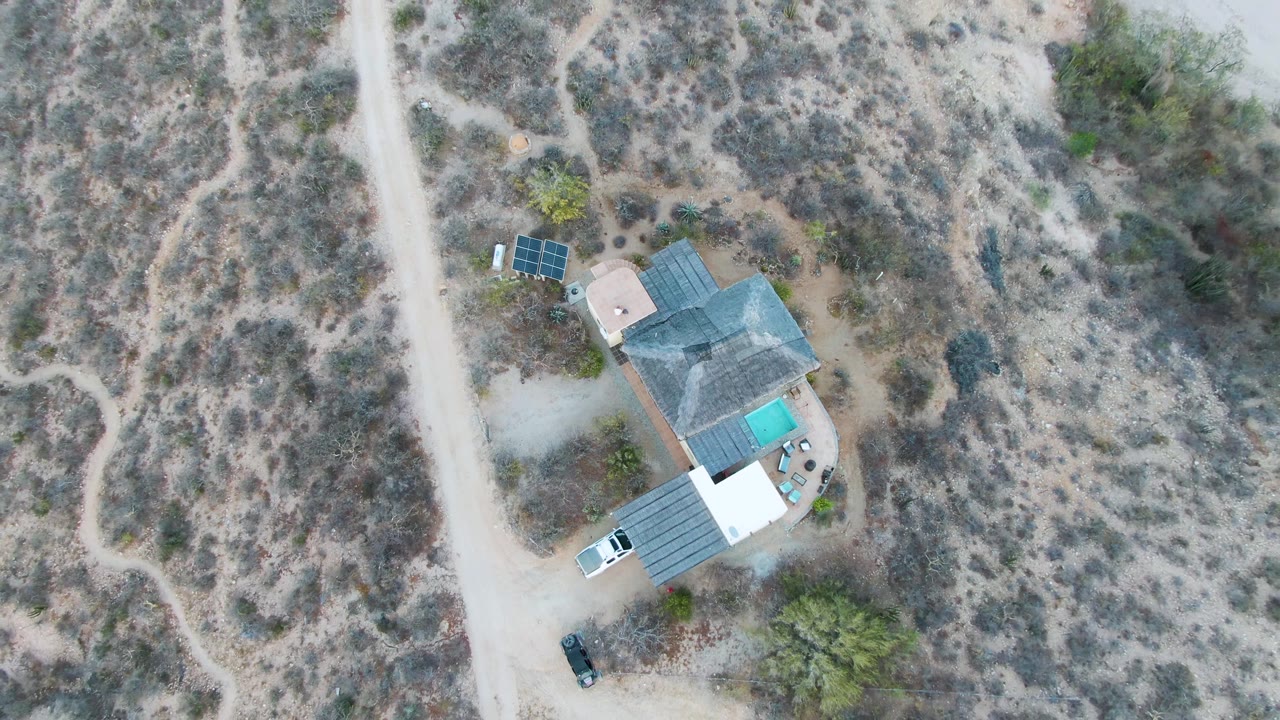 At 0:02, the top-down view shows the Cabo Pulmo desert home with the pool bright beside the roof, solar arrays nearby, pale driveway tracks, dry vegetation, and no visible people.