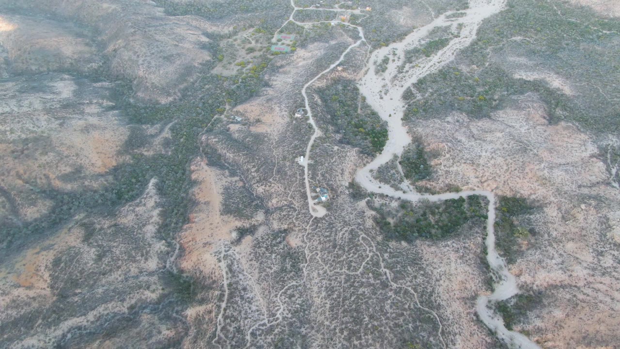 Aerial frame over Cabo Pulmo showing the winding dry waterway, branching dirt tracks, scrub patches, and a small vehicle near the wash with no visible people.