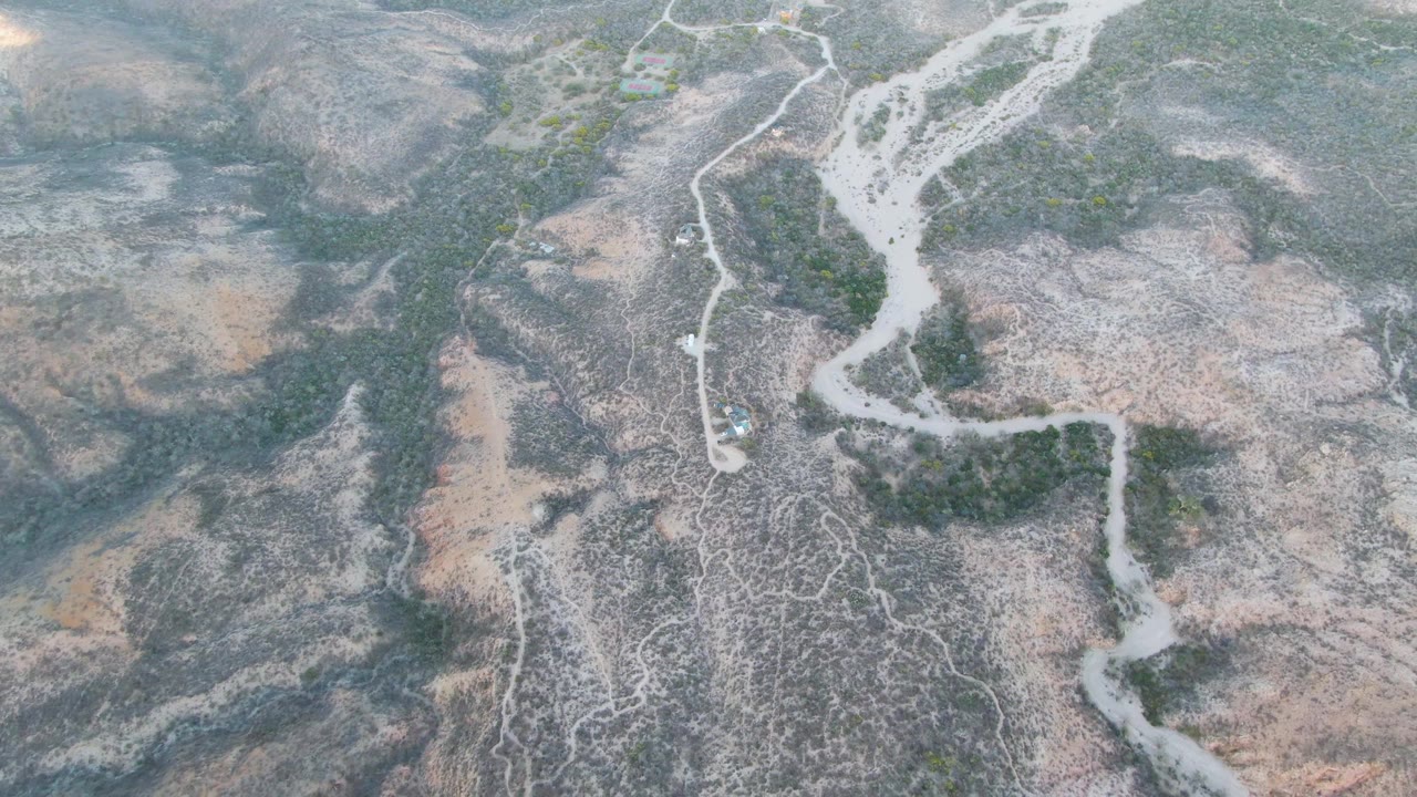Aerial frame over Cabo Pulmo showing a small vehicle near branching dirt paths at the edge of a pale dry riverbed, with scrub vegetation and no visible people.