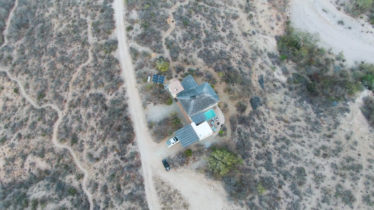 Overhead drone frame of a small house compound in Cabo Pulmo, Baja California Sur, with a pale dirt road on the left, scrubby desert terrain around it, and no visible people.