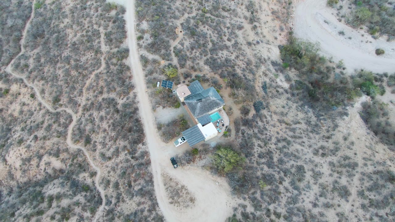 High drone frame over Cabo Pulmo, Baja California Sur, with the rural house near the lower left, a white vehicle by the road, winding footpaths or tracks across scrubland, and no visible people.