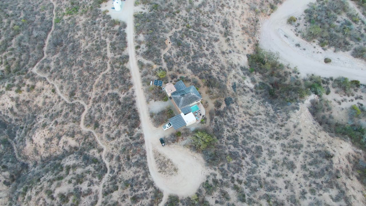 Overhead view in Cabo Pulmo, Baja California Sur, showing a pale dirt road beside a small house compound and a white trailer in dry scrubland, with no visible people.