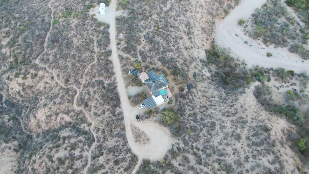 High drone view over Cabo Pulmo, Baja California Sur, showing the white trailer on a curving dirt road beside a compact house site set in scrub-covered slopes, with no visible people.