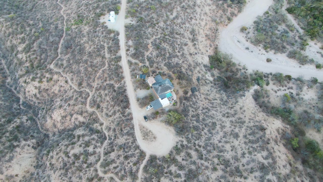 Overhead drone frame in Cabo Pulmo, Baja California Sur, showing a white vehicle on a pale dirt road near a small hillside house, scrub vegetation, and no visible people.