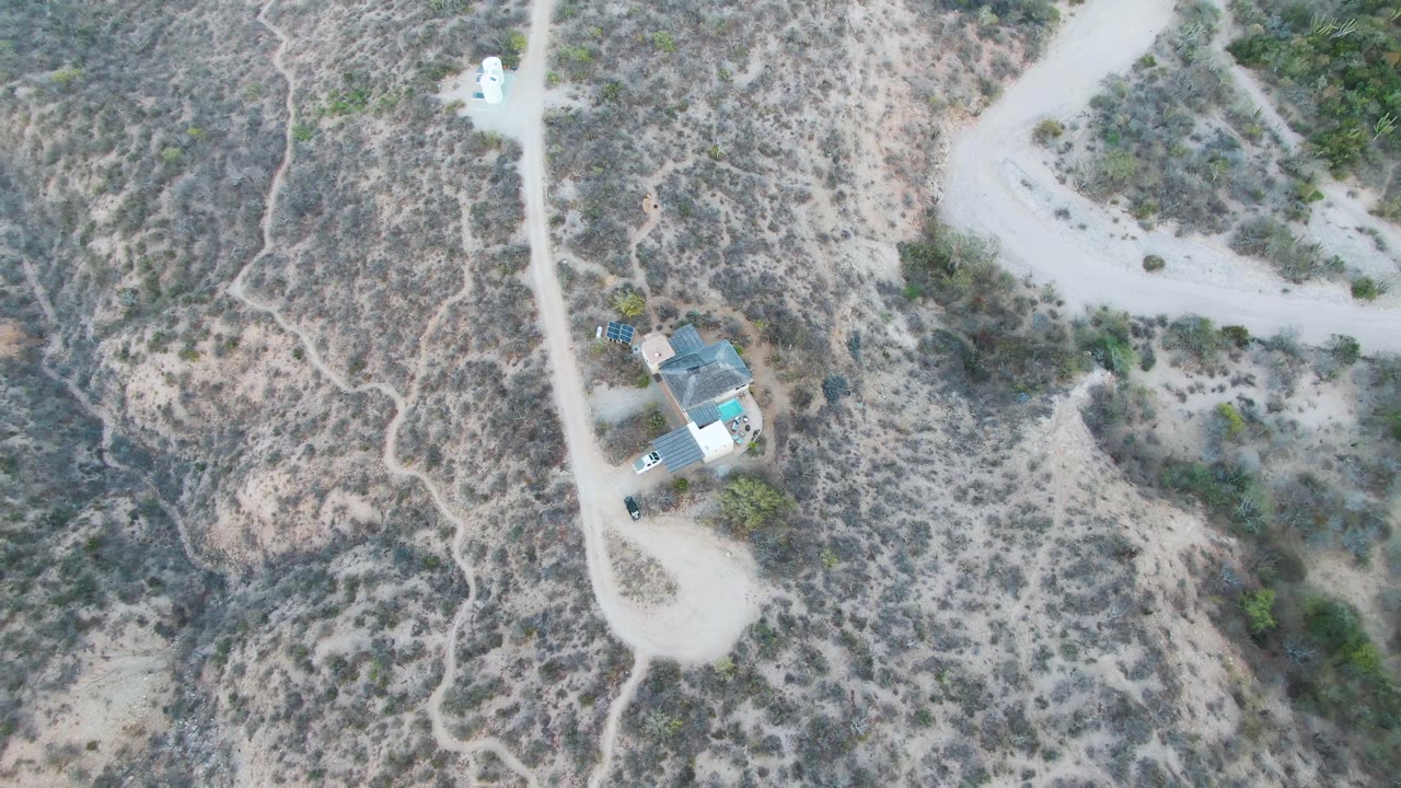 High overhead frame in Cabo Pulmo, Baja California Sur, with the white vehicle slightly farther along the road, the house near the lower left, dry slopes, and no visible people.