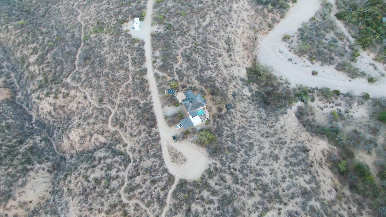 Top-down Cabo Pulmo frame showing the white vehicle on the dirt road, a driveway and house below it, scattered green shrubs, pale soil, and no visible people.