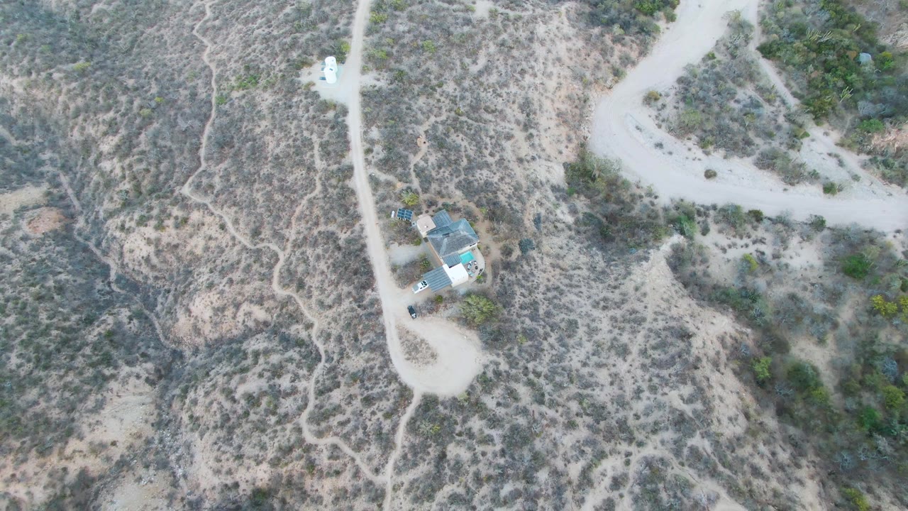 Overhead drone frame in Cabo Pulmo, Baja California Sur showing a pale dirt road, a small isolated house, dry scrubland, and no visible people.