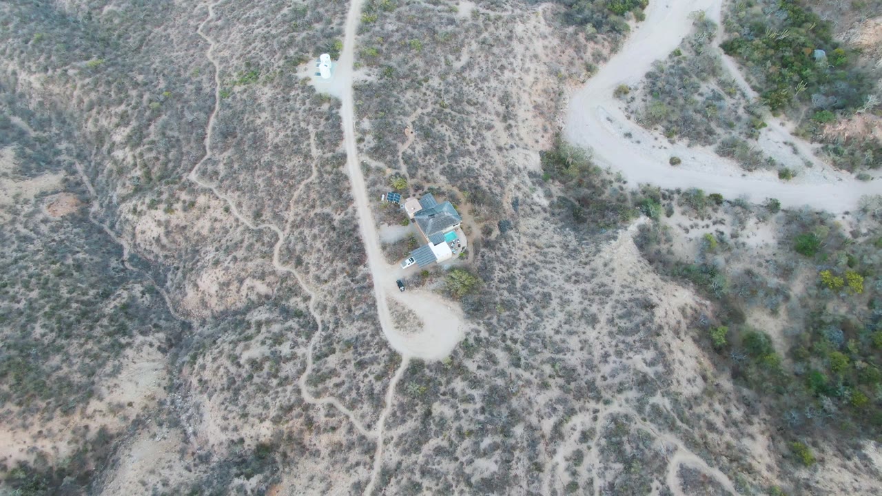 High overhead frame in Cabo Pulmo with the house lower in frame, faint footpaths across scrubland, pale roads, and no visible people.