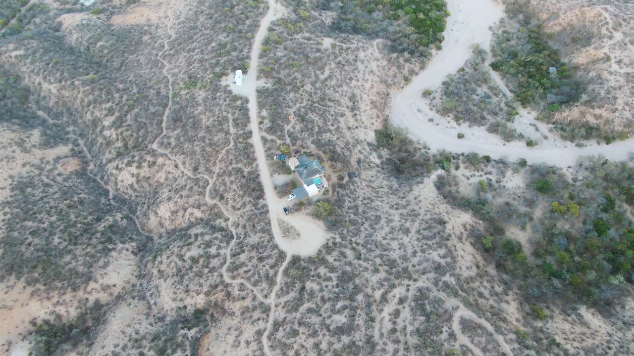Overhead view in Cabo Pulmo of a white vehicle on a pale dirt road near scrub-covered slopes and small buildings, with no visible people.
