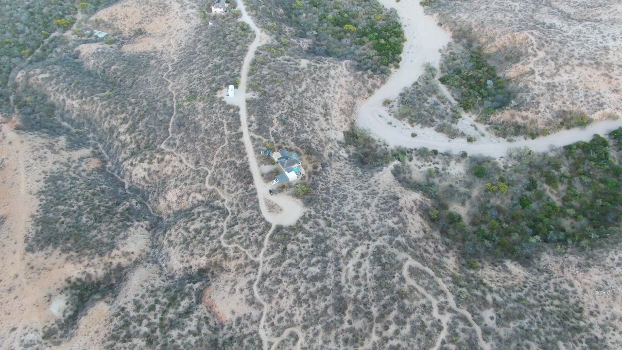 Overhead view of Cabo Pulmo arid hillside with pale winding dirt roads, patchy scrub, darker bushes, a small light-roofed structure near the lower left, and no visible people.