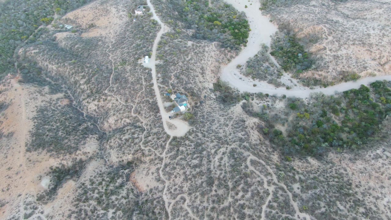 Overhead Cabo Pulmo frame with a pale road curving around a dense green bush area, dry brown slopes, scattered scrub, and no visible people.