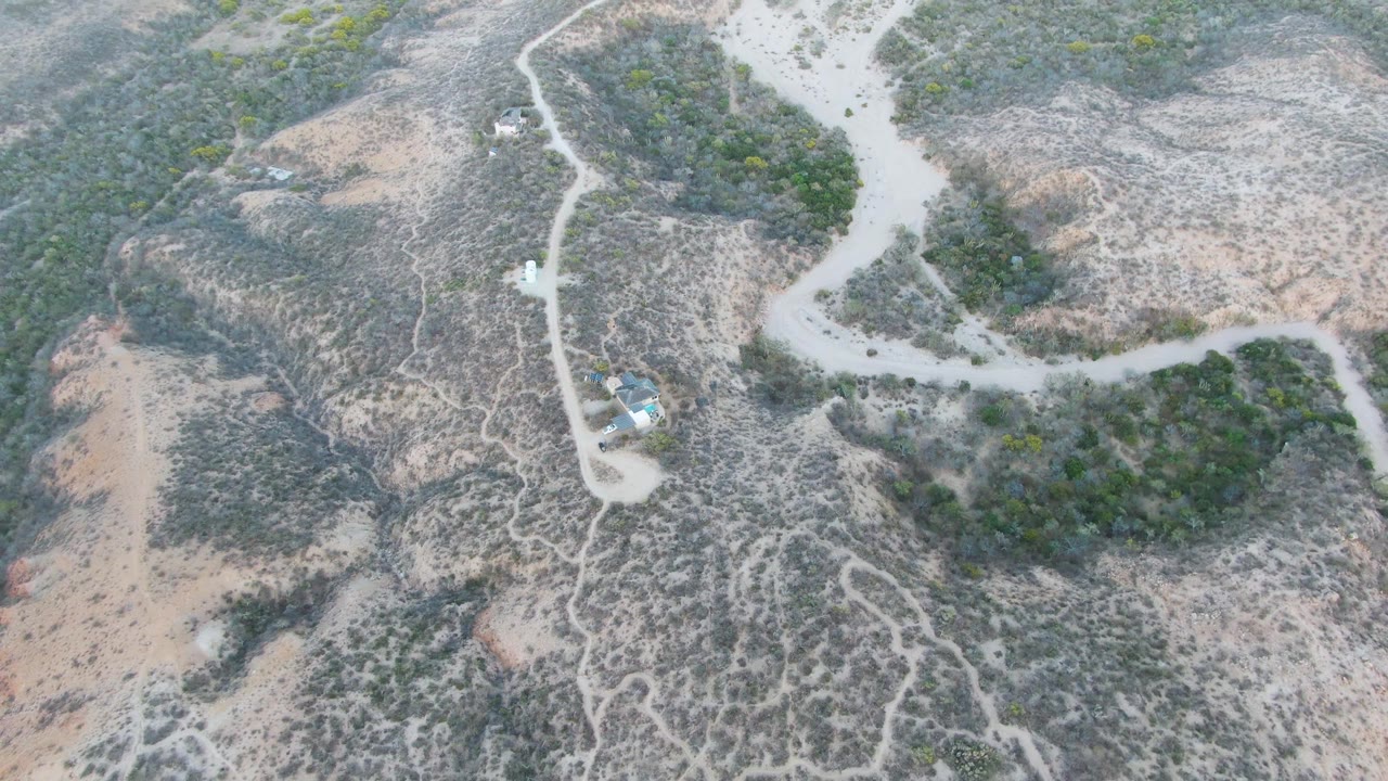 High aerial frame over Cabo Pulmo dry hills, showing a pale winding dirt road, scrub vegetation, a small vehicle near the upper left, and no visible people.