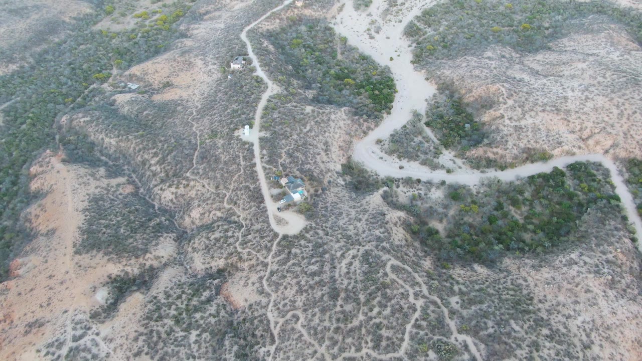 Aerial frame in Cabo Pulmo with the dirt road running from the upper edge toward a small structure near the lower left, surrounded by dry hills and no visible people.