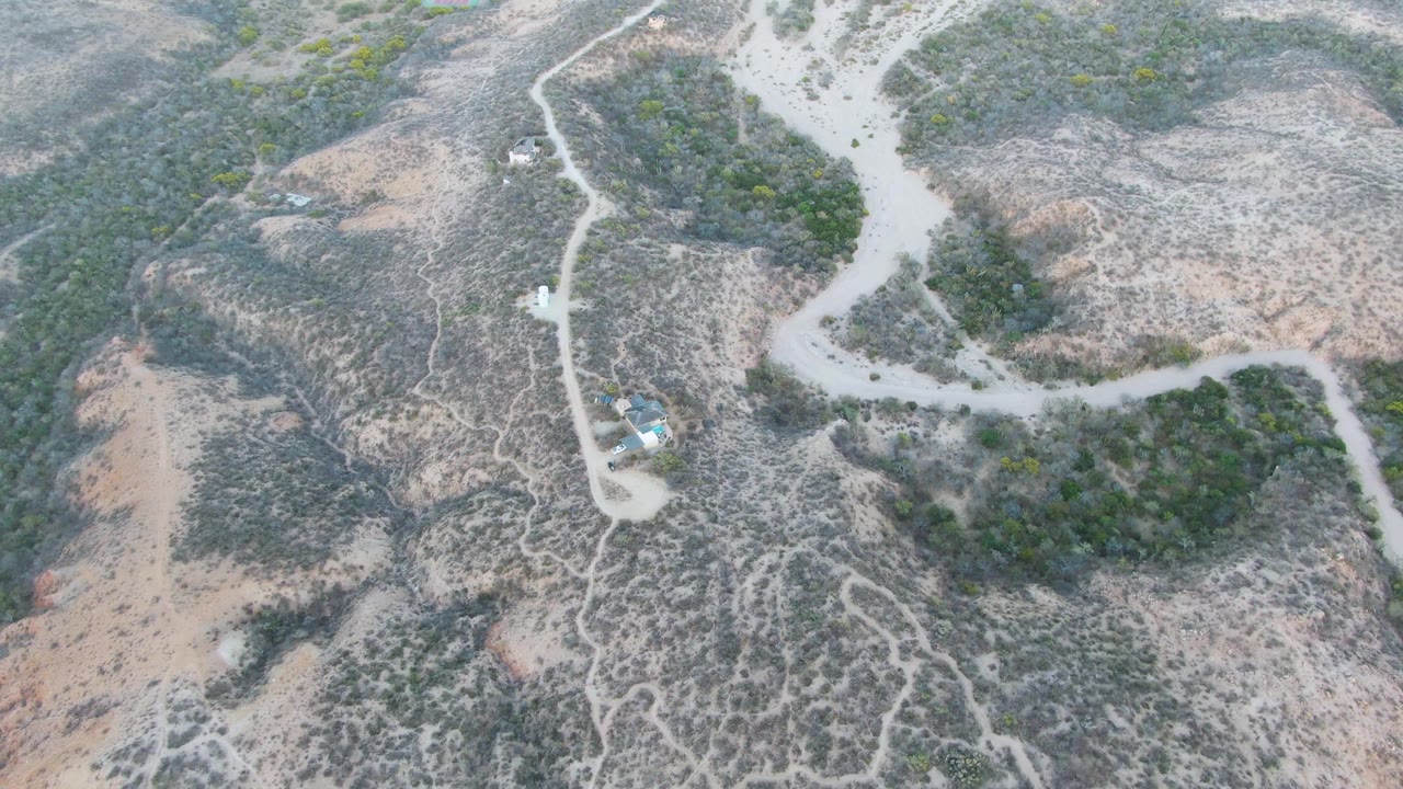 Cabo Pulmo aerial view with a pale road bending through scrub-covered hills, a tiny vehicle on the track, and no visible people in the open terrain.