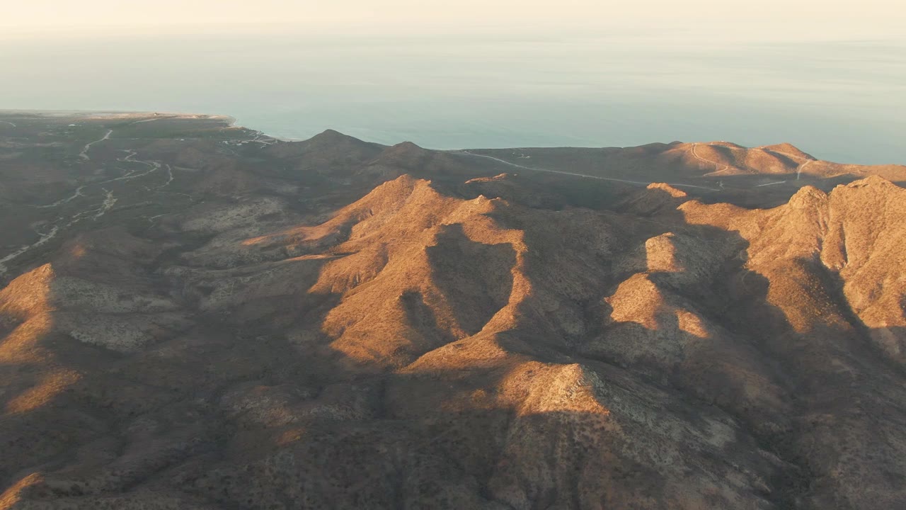 Aerial dusk view over brown and golden mountain ridges near Cabo Pulmo, Mexico, with the ocean in the distance and no visible people.