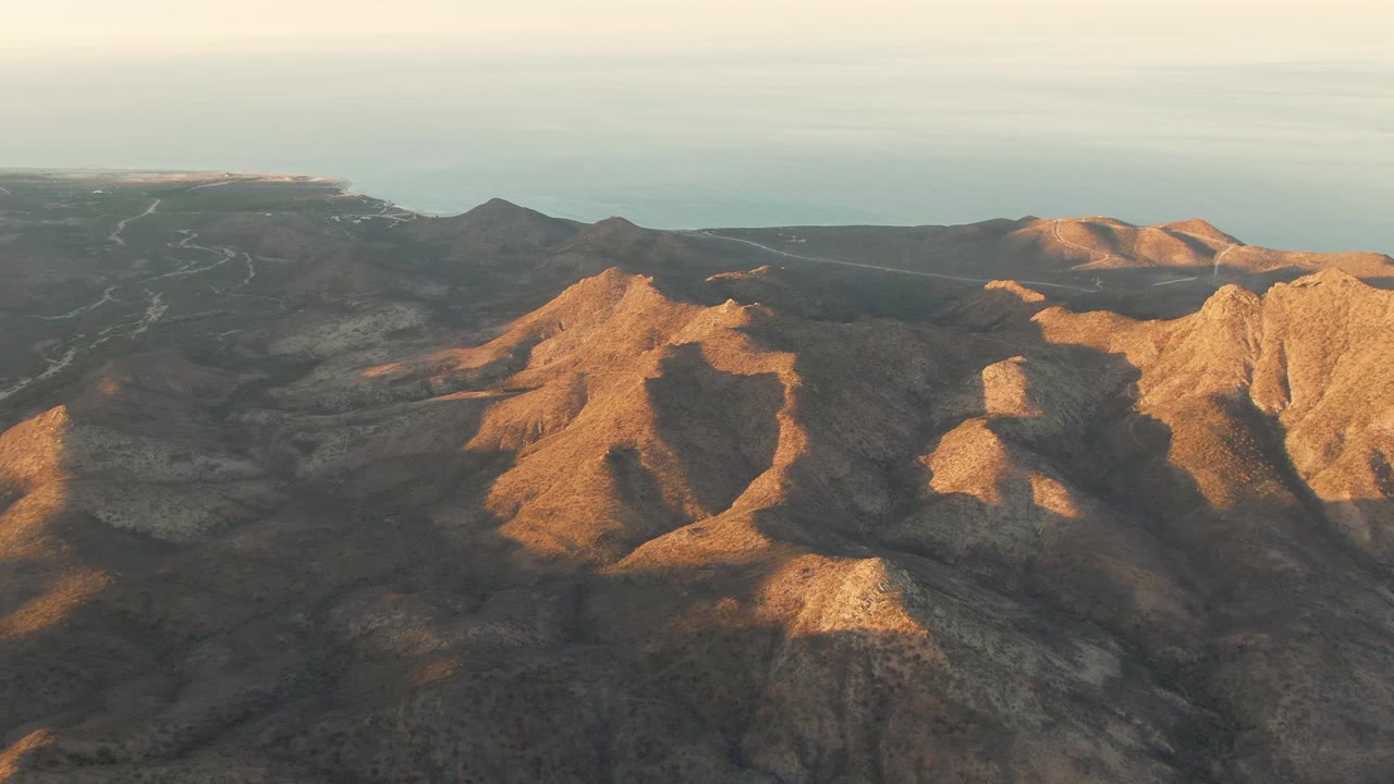 Aerial view of rugged East Cape mountain terrain with warm dusk light on the right-hand ridges, ocean beyond, and no visible people.