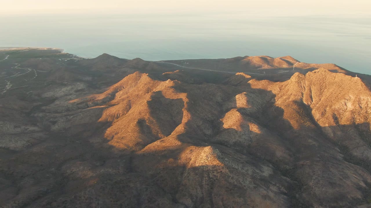 Aerial frame of Cabo Pulmo's rocky mountain terrain with a bright ridge at right, darker hills at left, and the ocean running across the background, no visible people.