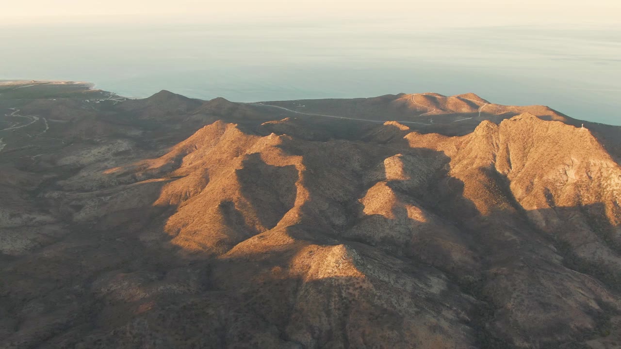 Aerial view of rugged Cabo Pulmo ridges with golden light on the central slopes, dark volcanic-looking hills behind them, and a soft sea horizon, no visible people.