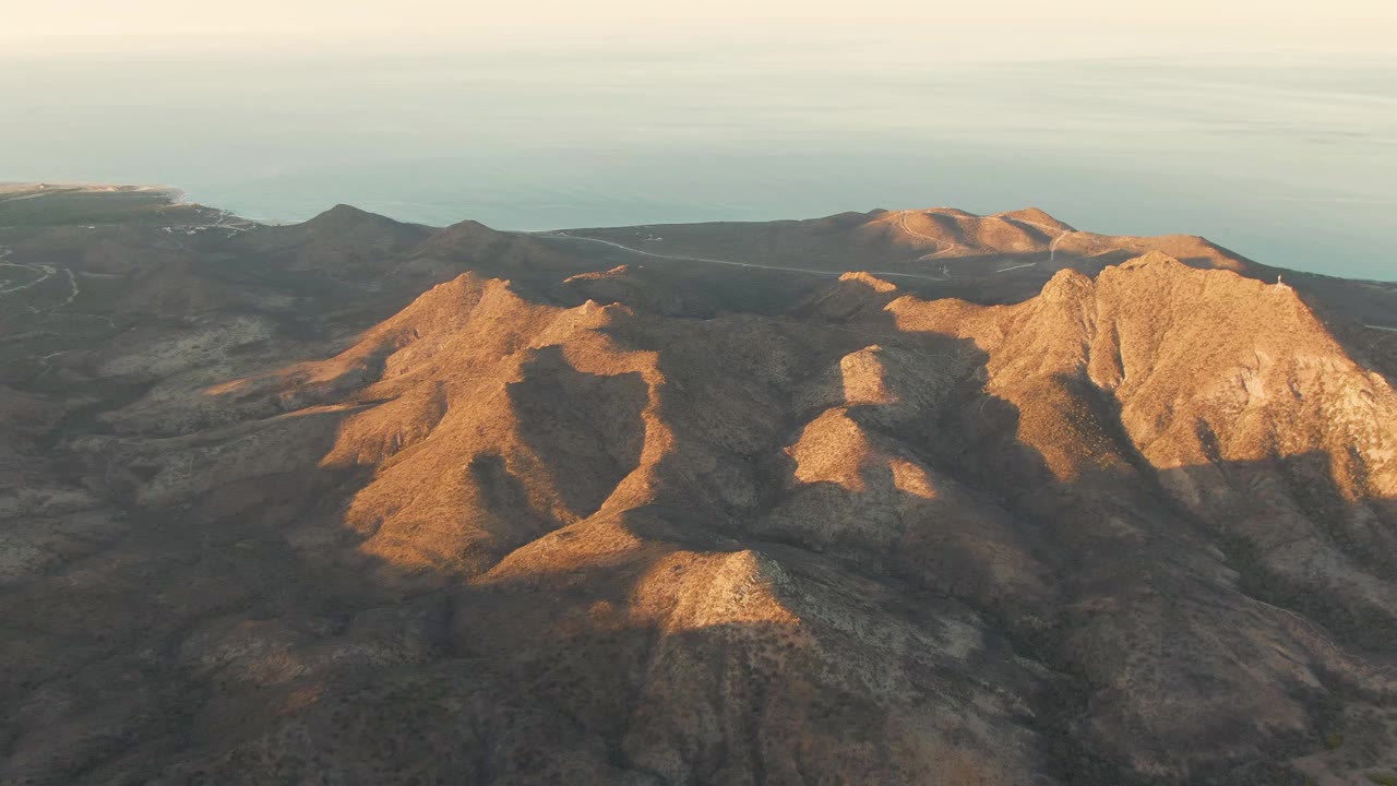 Aerial frame from Cabo Pulmo showing a broad rocky ridge in warm light, shadowed gullies across dry terrain, and the pale ocean beyond the hills, no visible people.