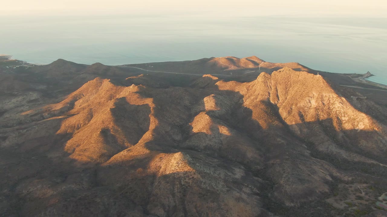 Sunlit ridges dominate the Cabo Pulmo aerial view, with the coastline and pale ocean visible beyond the dark foreground slopes and no visible people.