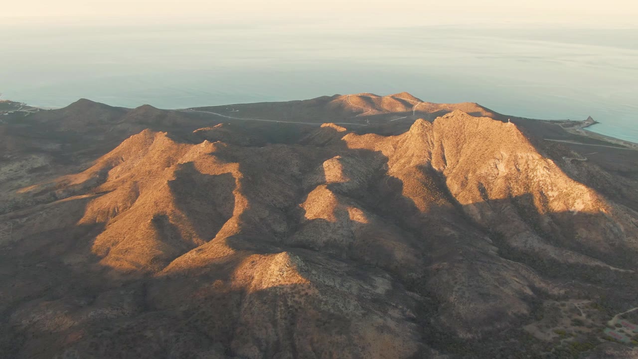 A wide drone view shows dry Cabo Pulmo mountain terrain, a bright rocky peak to the right, darker hills to the left, ocean along the horizon, and no visible people.