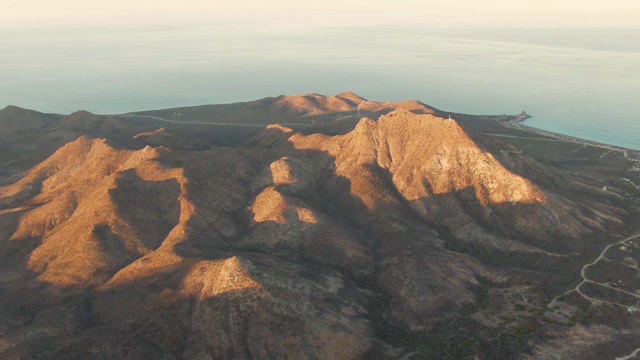 Aerial view over rocky Cabo Pulmo mountains with warm sun on tan ridges, pale ocean beyond, winding roads at right, and no visible people.