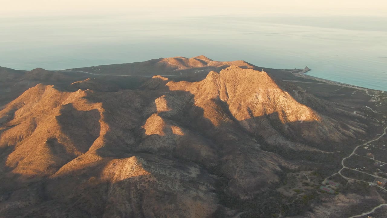 High aerial frame of Cabo Pulmo's rocky coastal mountains, with a brighter central ridge, the sea behind it, roads on the right slope, and no visible people.