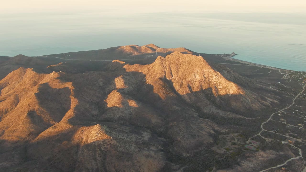 Aerial view of Cabo Pulmo coastal hills with golden light on the left ridges, a large pale rock face near center right, ocean across the background, and no visible people.